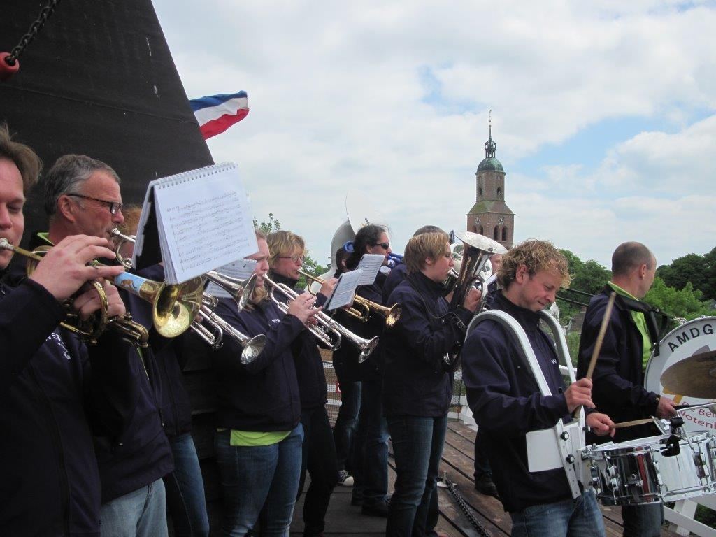Muzikanten van AMDG spelen op een molen, met de kerktoren van Beilen op de achtergrond